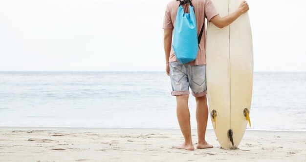 Colpo posteriore di giovane surfista caucasico a piedi nudi in piedi sulla spiaggia sabbiosa, appoggiandosi sulla sua tavola da surf e ammirando la bellezza e il potere dell'oceano di fronte a lui