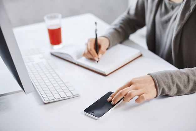 Colpo potato delle mani che annotano in blocco note e nello smartphone commovente. Dipendente che lavora in ufficio, controlla la posta via computer e beve succo di frutta fresco per aumentare l'energia. Le scadenze sono vicine