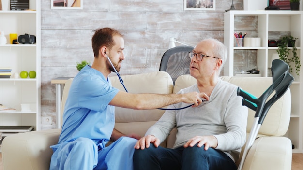 Colpo rivelatore di un giovane infermiere maschio che ascolta il battito cardiaco di un vecchio pensionato in una casa di cura luminosa e accogliente. Badante e assistente sociale