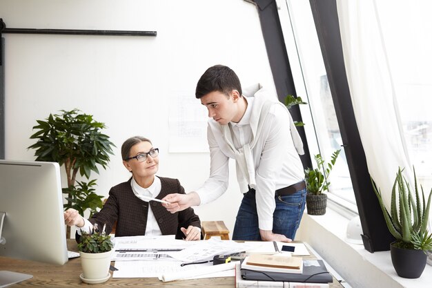 Colpo schietto di felice donna di mezza età architetto che lavora in ufficio con un giovane collega maschio che condivide idee creative e una nuova visione sul progetto di costruzione, puntando il dito sullo schermo del computer