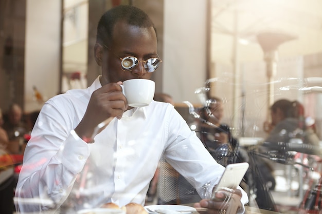 Colpo schietto di riuscito imprenditore nero elegante in occhiali da sole rotondi con cappuccino mattutino presso la caffetteria, utilizzando il telefono cellulare