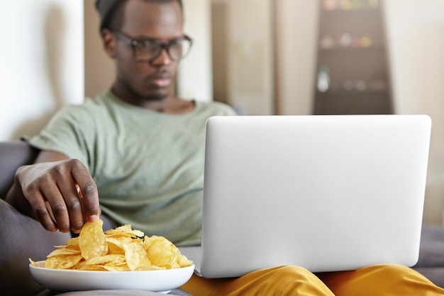 Colpo schietto di singolo maschio concentrato serio in cappello e vetri che si rilassano nel suo appartamento con il computer portatile e la zolla dei chip, rete praticante il surfing o guardare serie. Messa a fuoco selettiva sulla mano dell'uomo