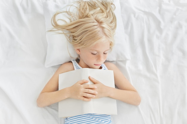 Colpo superiore del piccolo pisolino adorabile del bambino femminile a letto. Bambina carina con lentiggini e capelli biondi che dormono tranquilli con il libro aperto dopo aver letto storie interessanti, vedendo bei sogni d'oro