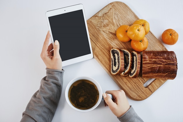 Colpo superiore delle mani femminili che tengono compressa digitale mentre facendo colazione. La donna intelligente aumenta l'energia con una tazza di tè e mangia i mandarini con la torta arrotolata, sentendosi benissimo la mattina d'inverno.