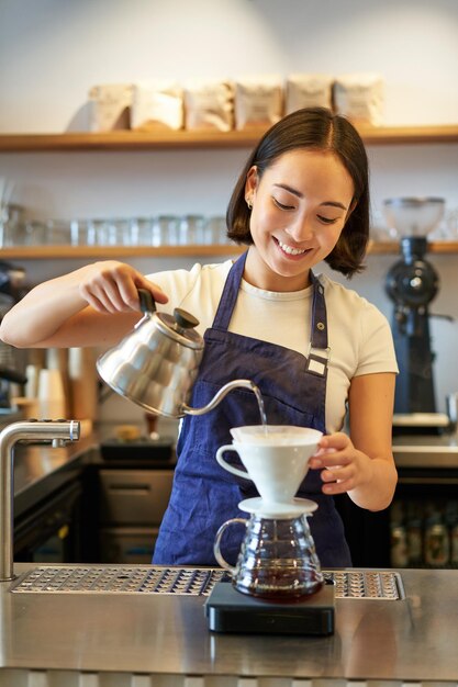 Colpo verticale del barista barista asiatico sorridente in grembiule blu che versa acqua con un piccolo bollitore brewi