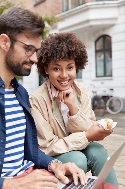 Colpo verticale del ragazzo barbuto concentrato lavora insieme al partner, keybroads sul computer portatile, mangia panino,