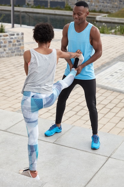 Colpo verticale dell'allenatore maschio aiuta il suo apprendista afroamericano a fare esercizi di stretching, stare all'aperto. La donna sportiva sta indietro, mostra una buona flessibilità, alza la gamba in alto, indossa scarpe da ginnastica.