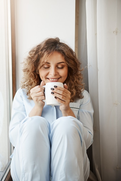 Colpo verticale della bellissima giovane donna caucasica con i capelli ricci seduto sul davanzale della finestra, tenendo la tazza di gustoso caffè, annusandolo con un sorriso compiaciuto e rilassato, avendo perfetta mattinata da solo