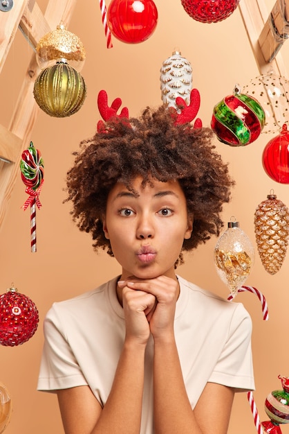 Colpo verticale della donna dai capelli ricci mantiene le labbra piegate e le mani sotto il mento guarda con espressione romantica alla telecamera vestita casualmente circondata da giocattoli di Natale ha un'atmosfera festosa. Celebrazione delle vacanze