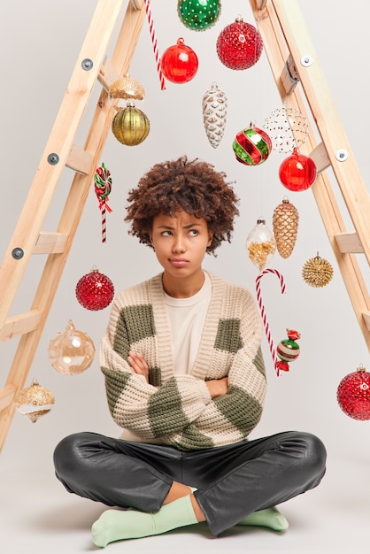 Colpo verticale della donna scontenta offesa si siede a gambe incrociate sul pavimento ha un'espressione infelice andando a decorare la casa con pose di palline di Capodanno