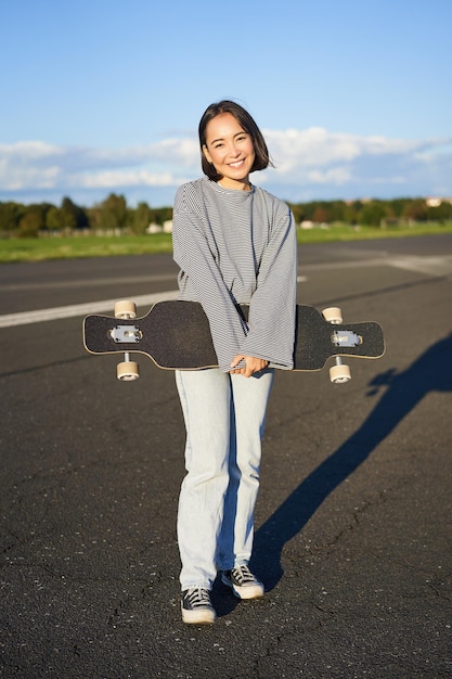 Colpo verticale della ragazza del pattinatore che posa con il longboard che gira sulla strada vuota nei sobborghi sorridente asiatico w