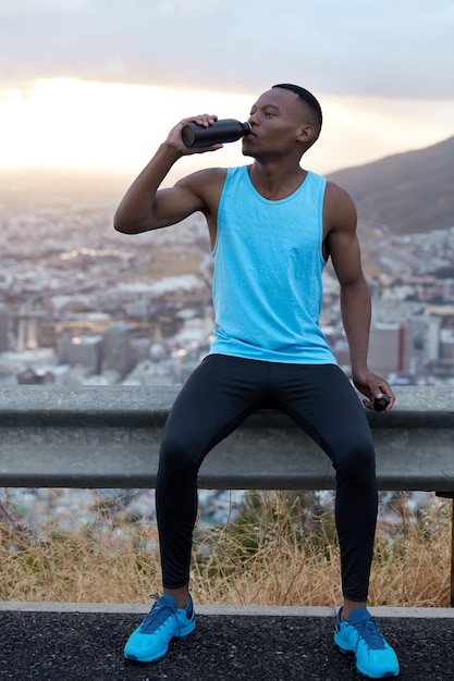 Colpo verticale di bell'uomo afroamericano in abiti sportivi beve l'acqua dopo l'allenamento fitness, si rinfresca con la bevanda, posa sulla collina di montagna, sente la stanchezza. Concetto di sport e ringiovanimento