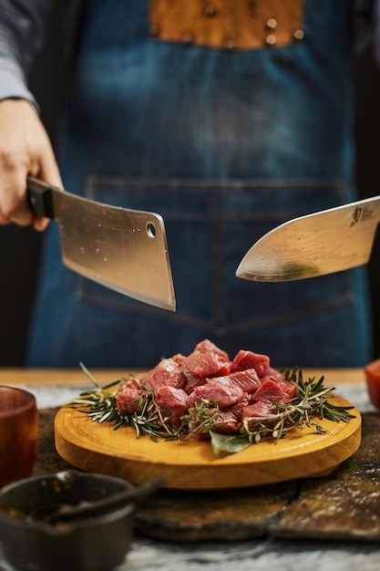 Colpo verticale di due coltelli nelle mani del cuoco sopra la carne cruda tagliata su un piatto di legno