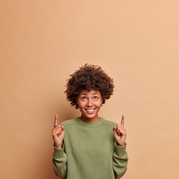 Colpo verticale di punti di modello femminile gioioso felice sopra con un ampio sorriso si sente lieto di mostrare lo spazio della copia per il tuo contenuto pubblicitario indossa un maglione casual isolato sul muro beige