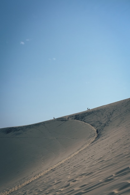 Colpo verticale di una duna di sabbia con persone che camminano in lontananza e un cielo blu chiaro