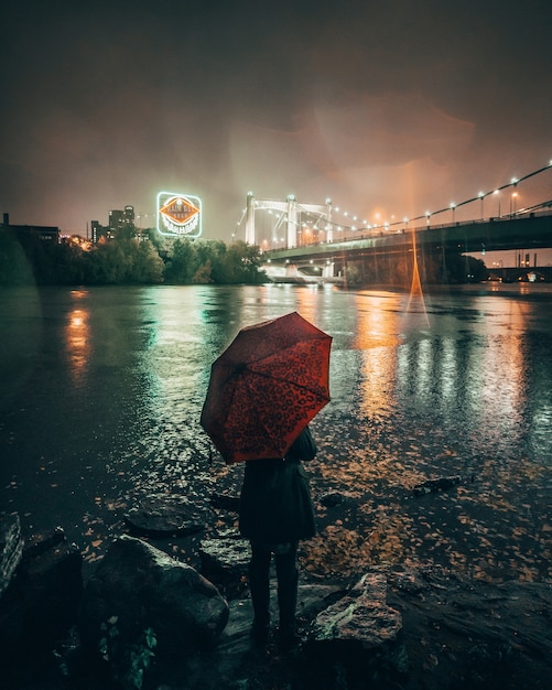 Colpo verticale di una femmina che tiene un ombrello rosso che sta vicino ad un lago nella città durante la notte