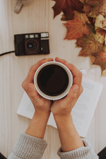 Colpo verticale di una persona in possesso di una tazza di caffè con una macchina fotografica e foglie in background