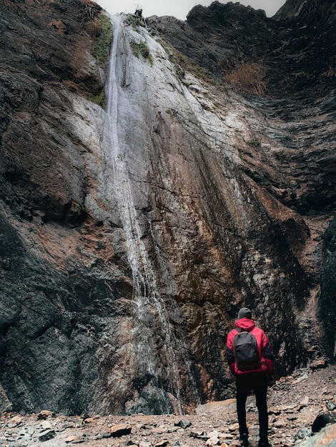 Colpo verticale di una persona in un cappotto rosso e uno zaino guardando un'alta scogliera con cascata