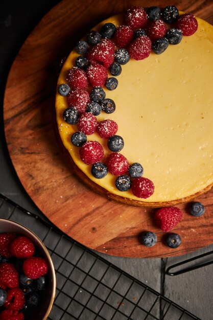 Colpo verticale di una torta di formaggio con frutti di bosco in cima su un piatto di legno con frutti di bosco sul lato