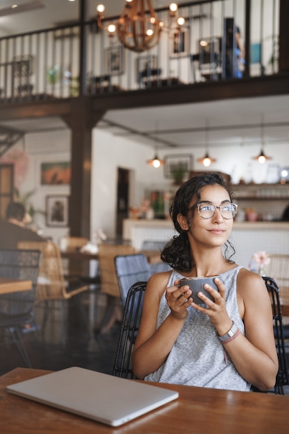 Colpo verticale dolce tenera donna urbana rilassata con gli occhiali godendo il momento seduto da solo nella caffetteria