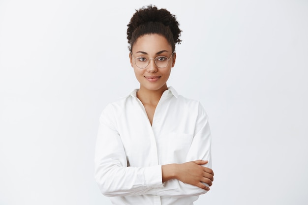 Come posso aiutarla. Affascinante donna afro-americana dall'aspetto amichevole in occhiali e camicia bianca, tenendo le mani incrociate sul petto. sorridendo educatamente, ascoltando il cliente, aspettando il cambiamento nel ristorante