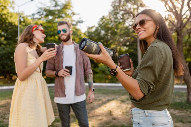 Compagnia di giovani hipster di amici che si divertono insieme nel parco sorridendo ascoltando musica su altoparlante wireless, stagione estiva