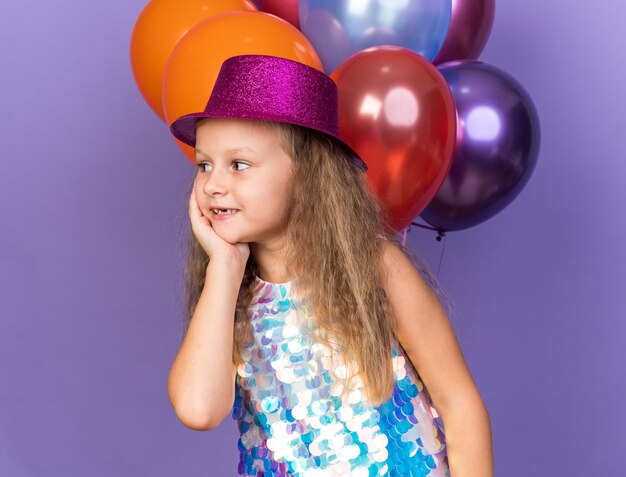 compiaciuta bambina bionda con cappello da festa viola che si mette la mano sul viso e guarda il lato in piedi con palloncini di elio isolati sulla parete viola con spazio di copia