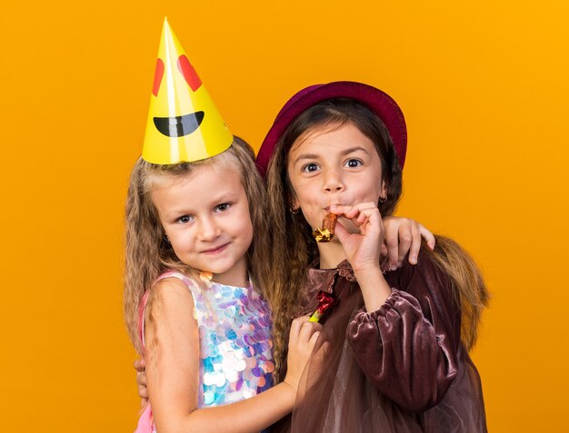 compiaciuta bimba bionda con cappello da festa che abbraccia gioiosa bambina caucasica con cappello da festa viola che soffia fischietto isolato sulla parete arancione con spazio di copia