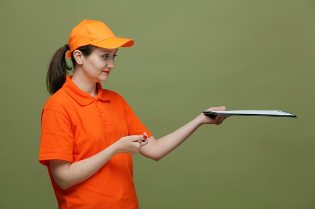 compiaciuta consegna di mezza età donna che indossa uniforme e cappuccio in piedi nella vista di profilo tenendo la penna allungando appunti guardando il lato isolato su sfondo verde oliva