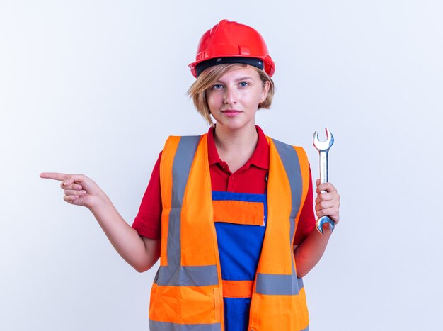 compiaciuta giovane donna costruttore in uniforme che tiene i punti chiave a forchetta sul lato isolato sul muro bianco con copia spazio