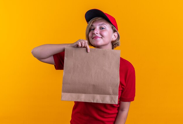 compiaciuta giovane ragazza delle consegne che indossa l'uniforme con il cappuccio che tiene il sacchetto di carta per alimenti isolato su parete arancione