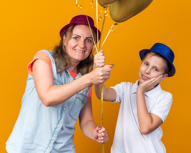 compiaciuto giovane ragazzo slavo con cappello da festa blu che mette la mano sul viso e tiene palloncini di elio con sua madre che indossa un cappello da festa viola isolato sulla parete arancione con spazio di copia