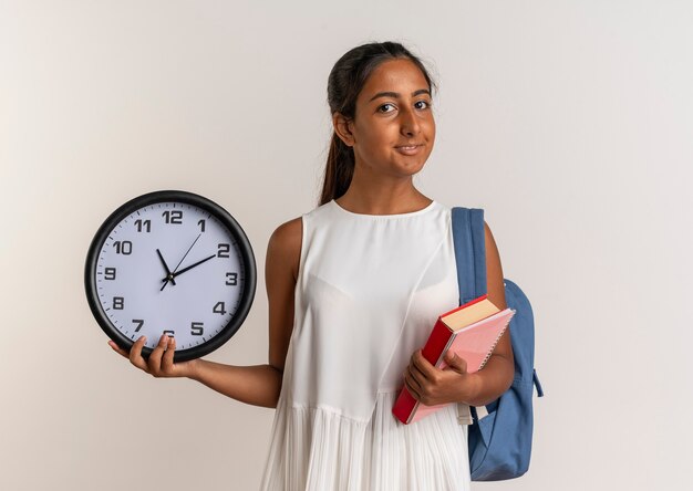 compiaciuto giovane studentessa che indossa la borsa indietro azienda libro con orologio da parete isolato su bianco