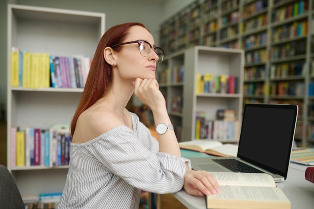 Comprensione. Giovane donna intelligente e pensierosa con occhiali con lunghi capelli rossi seduta davanti al computer portatile con libro aperto che guarda al lato in camera con scaffali