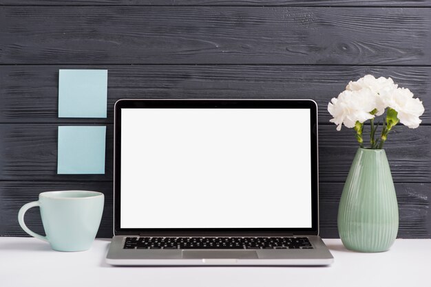 Computer portatile bianco dello schermo in bianco; vaso di fiori; tazza sullo scrittorio bianco contro la parete nera di legno