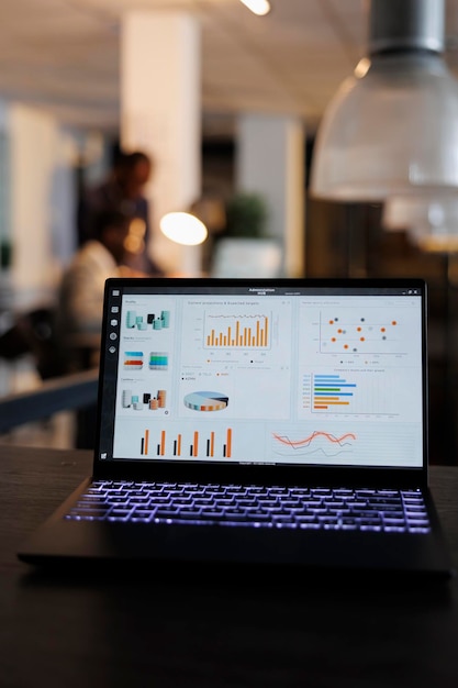 Computer portatile con grafici delle statistiche di marketing sullo schermo, in piedi sul tavolo nell'ufficio di avvio, colleghi che discutono della strategia aziendale in background. Dipendenti che fanno gli straordinari nell'ufficio di avvio