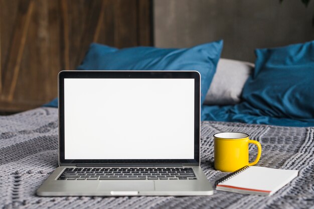 Computer portatile con lo schermo bianco in bianco vicino alla tazza e al blocco note a spirale sul letto