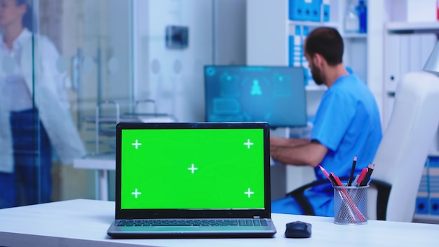 Computer portatile con schermo sostituibile nell'armadietto dell'ospedale, medico che indossa un cappotto che arriva in clinica e infermiere che scrive prescrizione. Notebook con schermo verde in clinica medica.