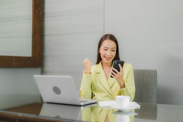Computer portatile del computer di uso della bella giovane donna asiatica del ritratto con il telefono cellulare astuto sul tavolo di lavoro alla stanza interna
