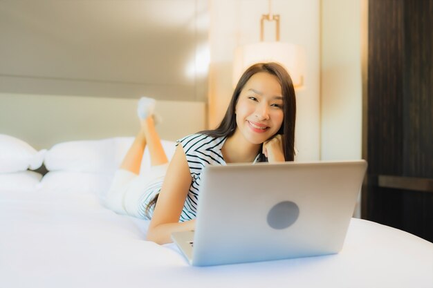 Computer portatile del computer di uso della bella giovane donna asiatica del ritratto sul letto nell'interno della camera da letto