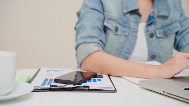 Computer portatile funzionante della bella giovane donna asiatica sorridente sullo scrittorio in salone a casa. Finanza e calcolatore del documento del taccuino di scrittura della donna di affari dell&#39;Asia in Ministero degli Interni.