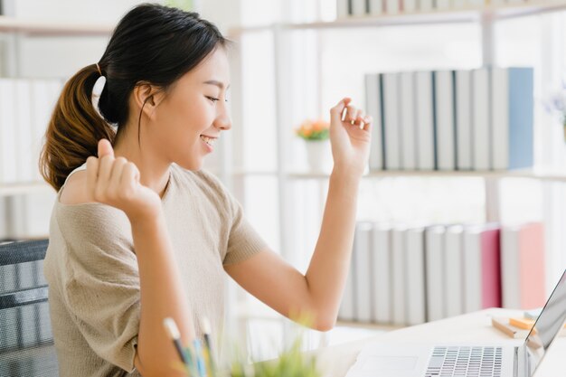 Computer portatile funzionante della bella giovane donna asiatica sorridente sullo scrittorio in salone a casa