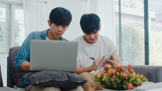 Computer portatile funzionante delle giovani coppie gay asiatiche a casa moderna. L'Asia LGBTQ + gli uomini si rilassano felici usando il computer e analizzando insieme le loro finanze in Internet mentre si trovano il divano nel soggiorno di casa.