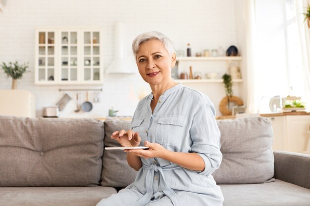 Comunicazione, dispositivi elettronici e networking. Donna di sessant'anni in pensione elegante moderna che indossa un abito blu lungo, navigare in internet utilizzando la tavoletta digitale, fare acquisti online o leggere e-book