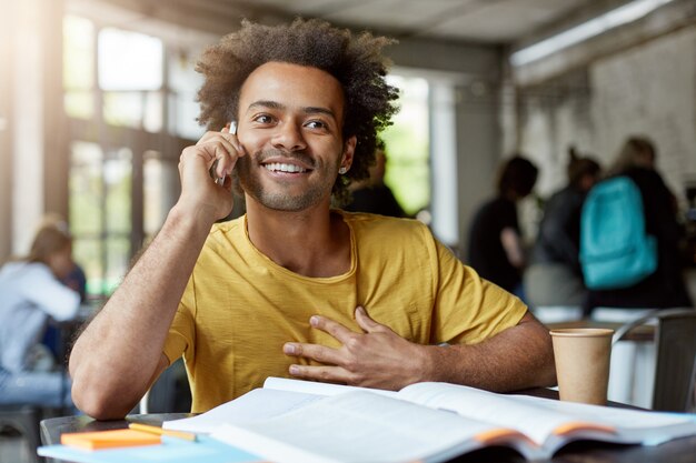 Comunicazione, istruzione e tecnologia moderna. Attraente studente di carnagione scura positivo con taglio di capelli afro seduto al tavolo del bar con libri di testo e godersi la conversazione telefonica