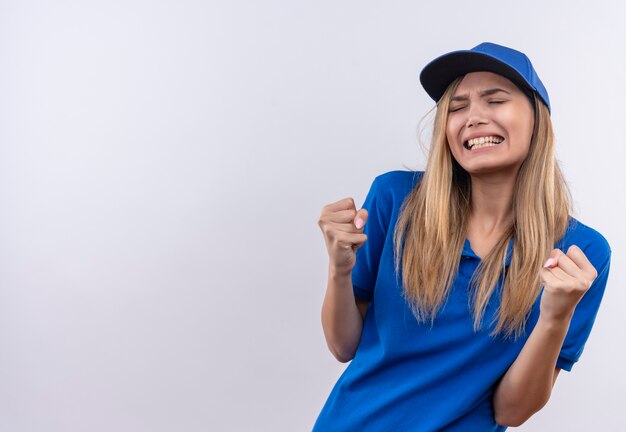 Con gli occhi chiusi allegra giovane ragazza di consegna che indossa l'uniforme blu e il cappuccio che mostra sì gesto isolato su bianco