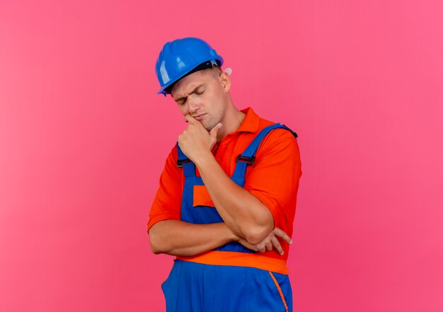 Con gli occhi chiusi che pensa giovane costruttore maschio che indossa l'uniforme e il casco di sicurezza che mette la mano sotto il mento sul colore rosa