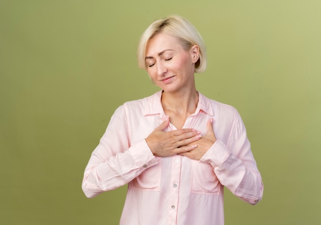 Con gli occhi chiusi compiaciuta giovane donna bionda slava che mette le mani sul cuore isolato sulla parete verde oliva