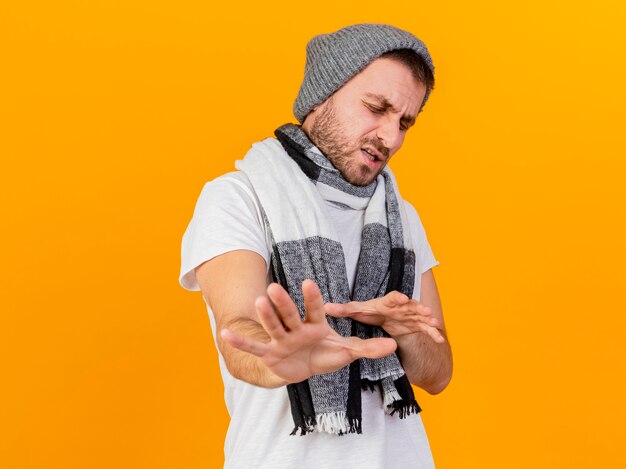 Con gli occhi chiusi dispiaciuto giovane uomo malato che indossa cappello invernale e sciarpa tendendo le mani alla telecamera isolata su giallo