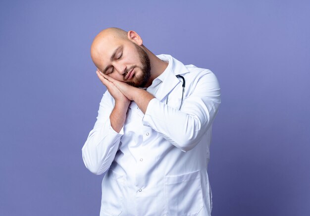 Con gli occhi chiusi giovane medico maschio calvo che indossa veste medica e stetoscopio che mostra il gesto del sonno isolato su priorità bassa blu